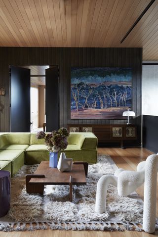 Image of a modern-retro living room. The walls are dark gray wood, and the ceiling and floors are natural wood. There is a fuzzy rug and a bright green sofa on top of it. The coffee table is in a futuristic shape. There is a white, boucle accent chair in a futuristic, abstract shape as well.