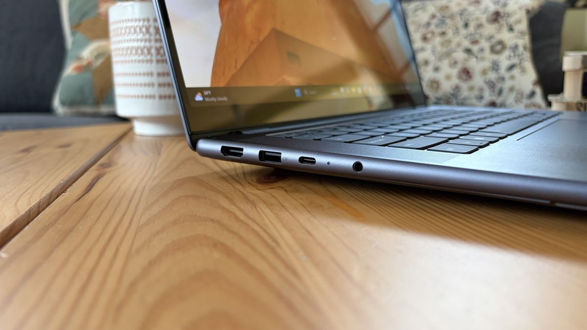 Close-up of a slim laptop on a wooden table, showing its keyboard and side ports; a patterned mug and cushions create a cozy workspace.