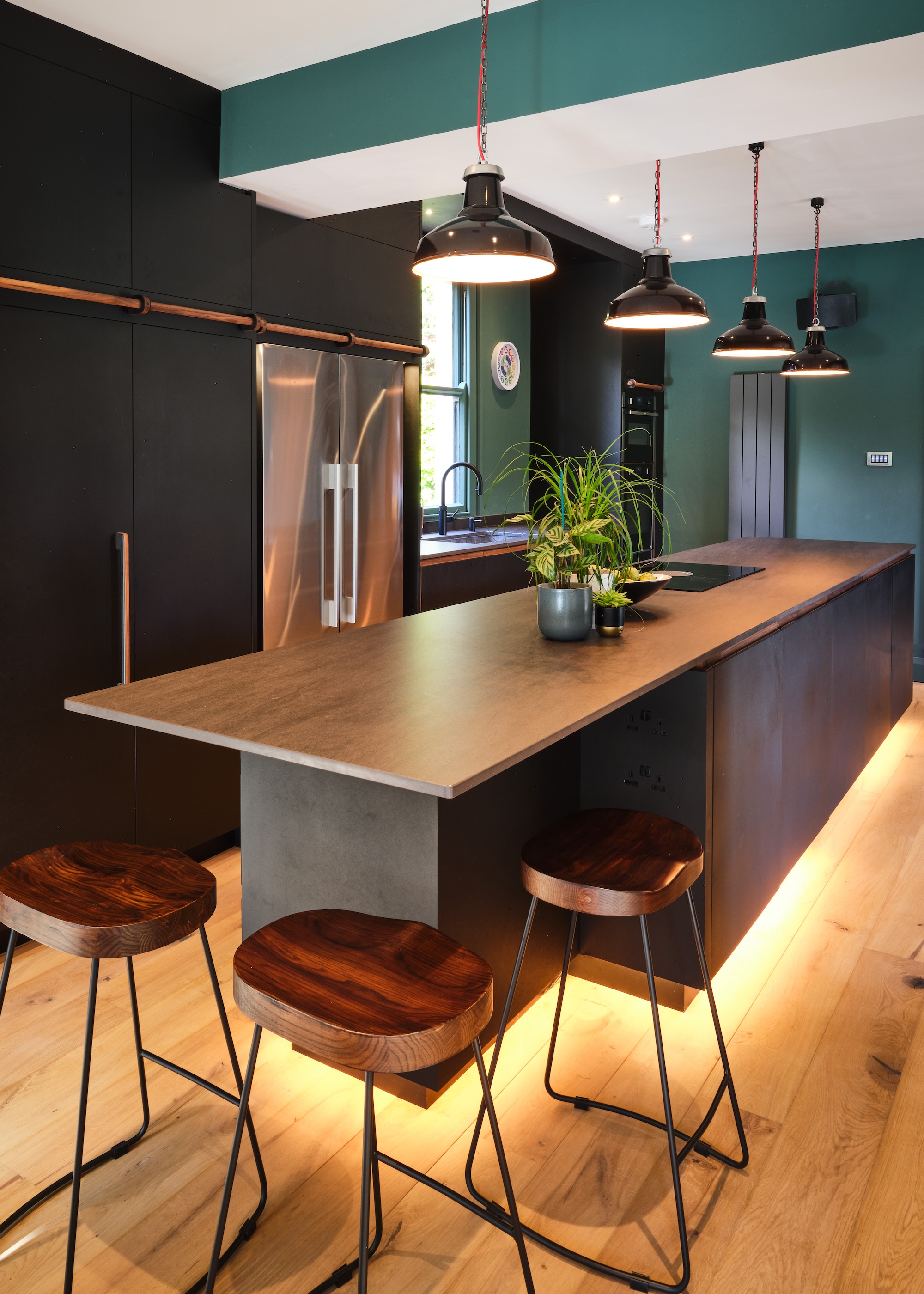 A large kitchen island with pendant lights hanging above as well as bar stools placed next to the kitchen island. There is also a view of a large double door fridge and floor to ceiling kitchen cabinets