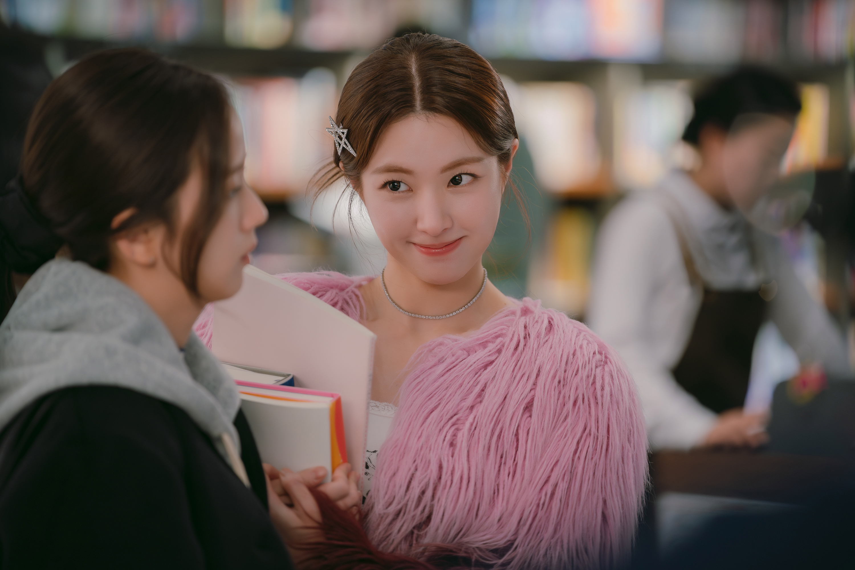 A woman (Ha Young as Lee Ji-yeon, middle) wears a pink feathered jacket while holding a stack of books and looking at another woman (Jisoo as Seo Mi-rae, left), who's wearing a hoodie and looking away. A female bookstore employee is seen in the blurred background, in a still from the Netflix K-drama 'Boyfriend on Demand.'