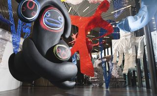 Alternative view of Charles Jeffrey's 'The Come Up' exhibition featuring large black figures with colourful faces, a white 3D hand, a red silhouette of a body, a brown and white arch and other blue sculptures