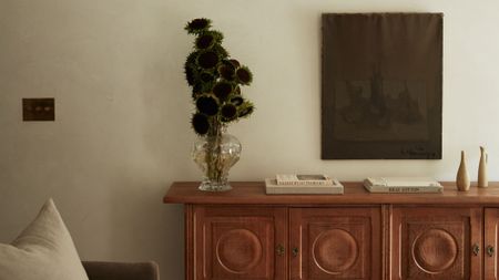 Warm minimalist living room featuring warm white plaster walls, a mid-century wooden sideboard, monochromatic still life painting, and an irregular vessel of sunflowers.