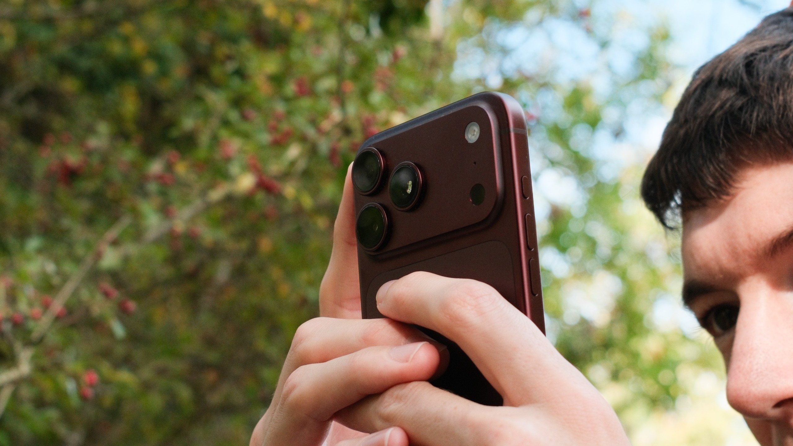 Close-up of a burgundy iPhone 18 Pro Max being held outdoors, showing the raised triple-camera module.