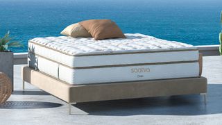 Image shows the Saatva Classic luxury innerspring hybrid on a beige bedframe overlooking a blue ocean on a sunny day