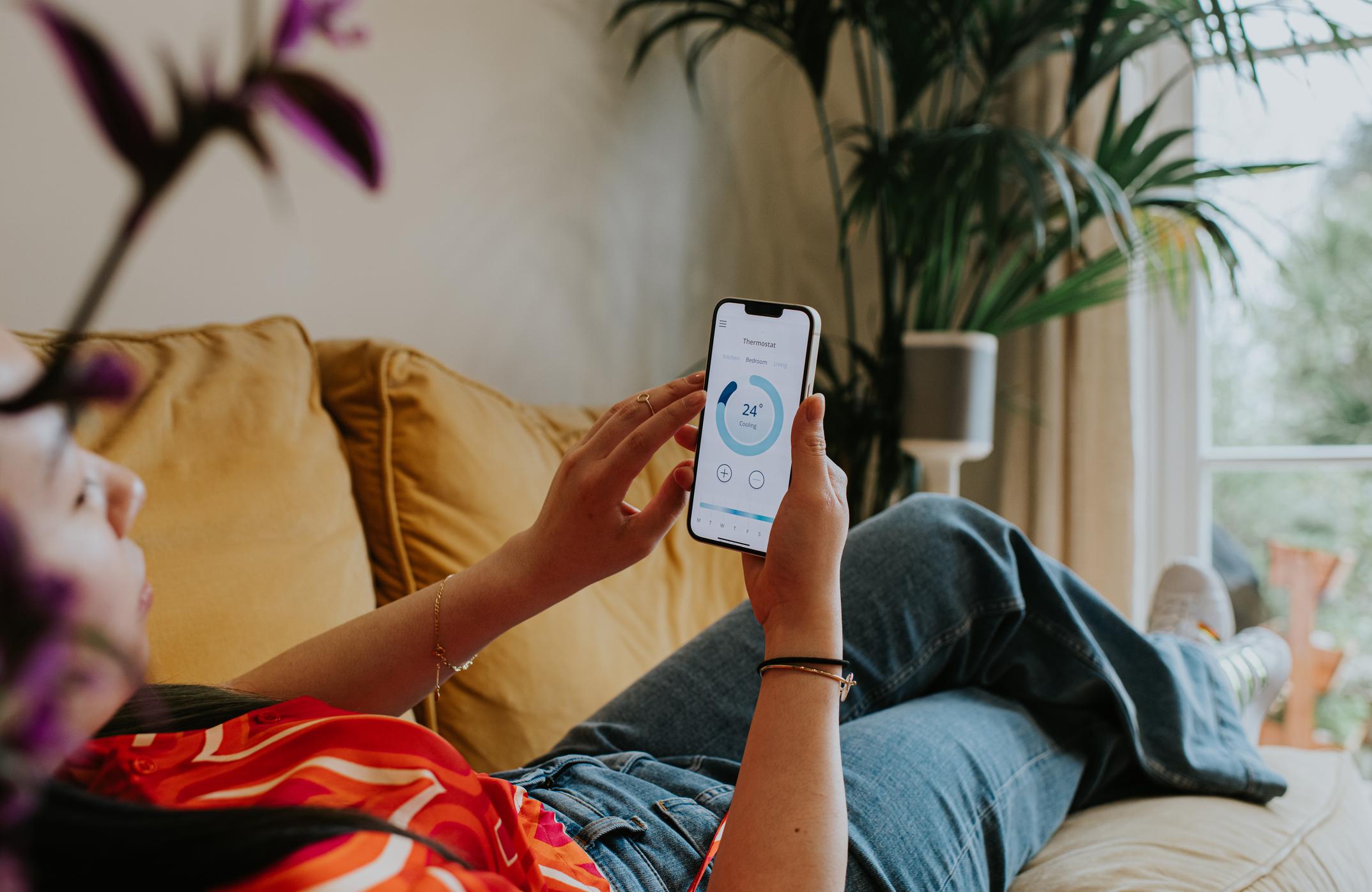  A woman relaxing on the sofa using a thermostat app on her smart phone. 