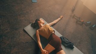 Woman lying on her back on yoga mat in studio with arm outstretched and leg over body for hip stretch
