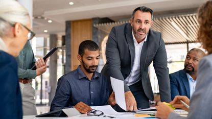 A stern-looking man presides over an office meeting where others look concerned.