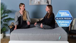 The image shows two women sat on a grey mattress, which is placed on a white bed frame in a blue bedroom. Ont the top of the image a badge reads 'Tom's Guide Black Friday Mattress Deals'
