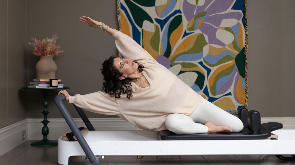 Rebecca Daodun, Pilates instructor, exercises on a Pilates reformer machine. Her knees are on the base of the machine, her right hand on the bar at the top, and her left hand reaching up and over her body in a stretch. 