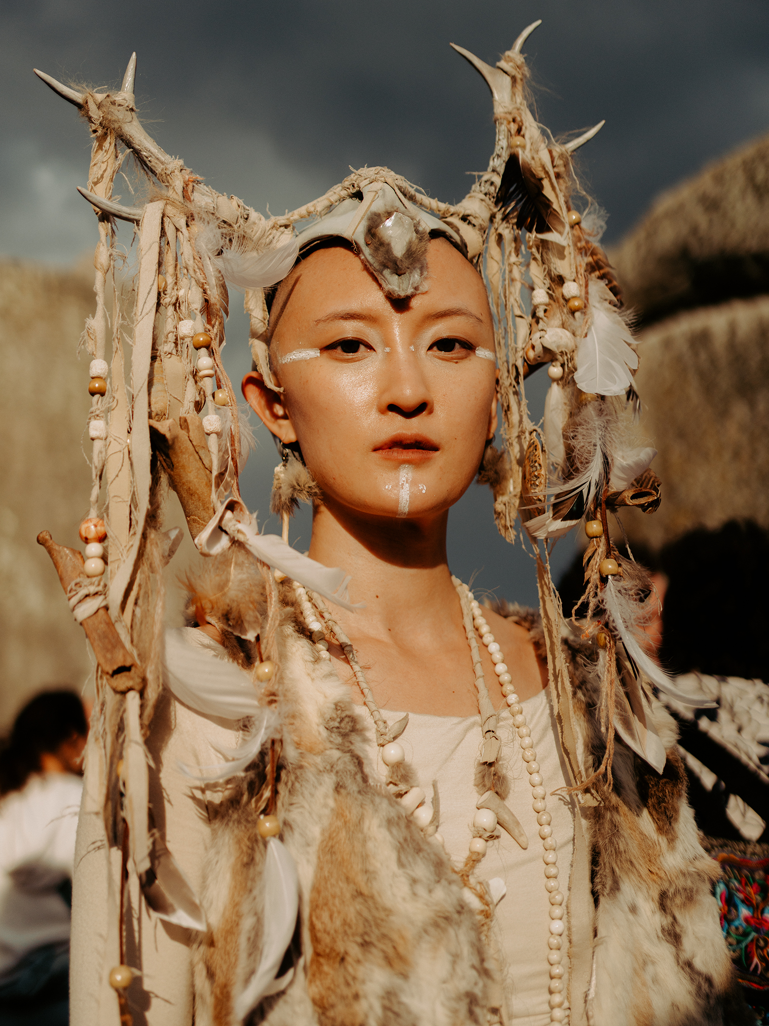 A person in traditional attire adorned with feathers, beads, and fur, set against a dramatic sky