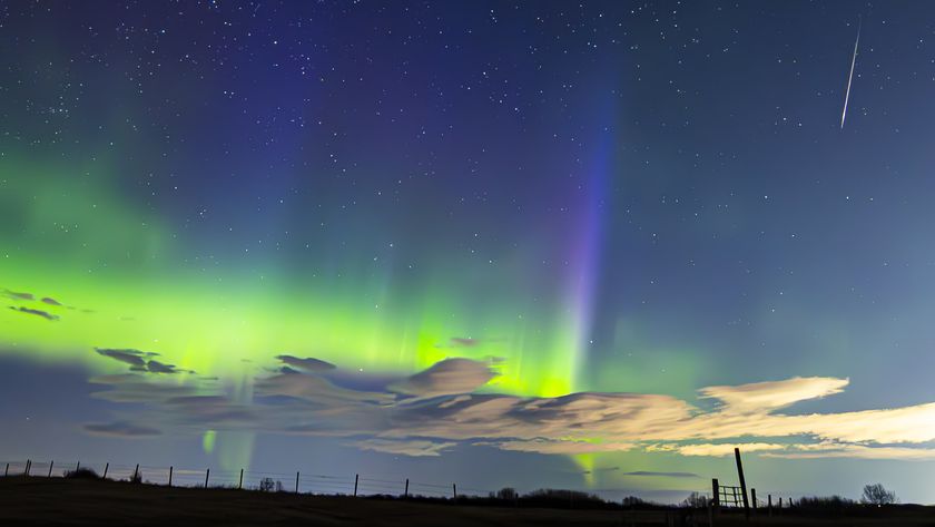 Blue and green aurora shine in a starry night sky above a dark field as a meteor streaks earthward at the right of the photo.