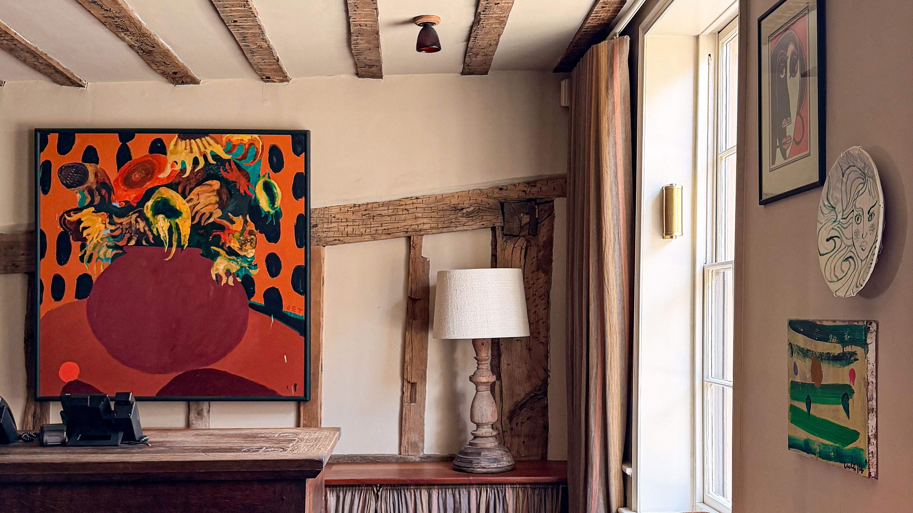 Room with wooden ceiling and wall beams, table lamp on wooden sideboard and walls with artwork and decorative plate