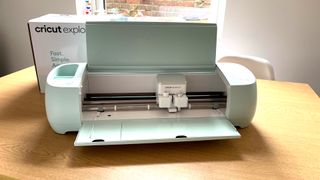 The best Cricut machines; a mint green craft cutting machine on a wooden table with its lid open, a box and window are in the background