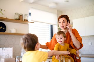 Mother with small children indoors in kitchen in the morning at home, using telephone. - stock photo