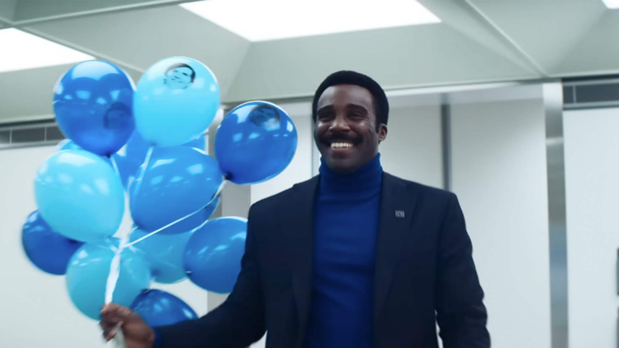 Tramell Tillman in a blue turtleneck and blue jacket, holding a bunch of blue balloons in Severance