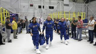 Three astronauts in blue spacesuits walking while others clap for them