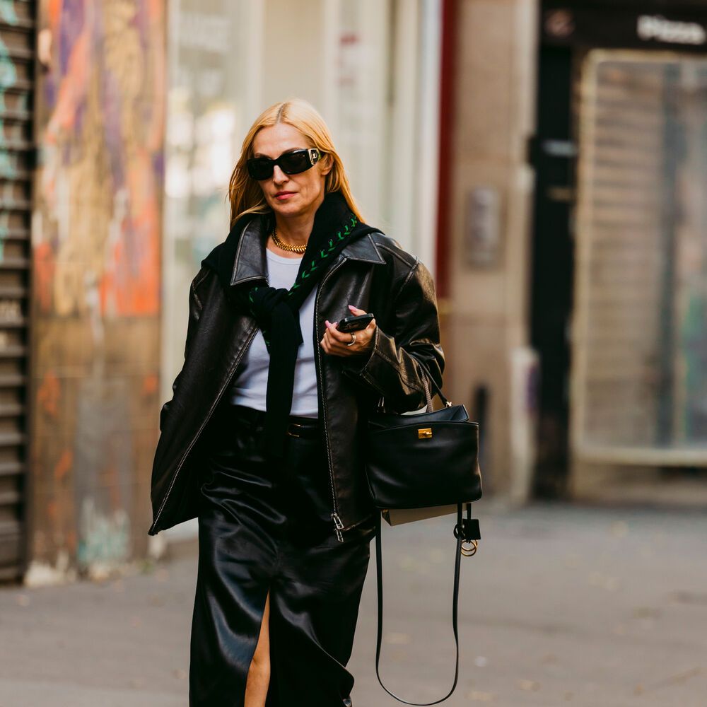 Woman wears black leather jacket, skirt, and handbag. She wears a lighter blue or white top with a black sweater tied around her neck. She has pointed toe heels with a lot of metal hardware on them. 