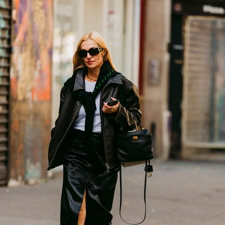 Woman wears black leather jacket, skirt, and handbag. She wears a lighter blue or white top with a black sweater tied around her neck. She has pointed toe heels with a lot of metal hardware on them. 