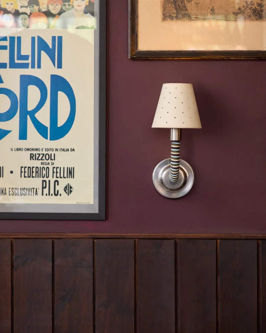 Image of a wall that is plum purple with dark wood paneling on the lower half. There are two framed pictures in view and a small silver sconce light with a beige pinhole lampshade.