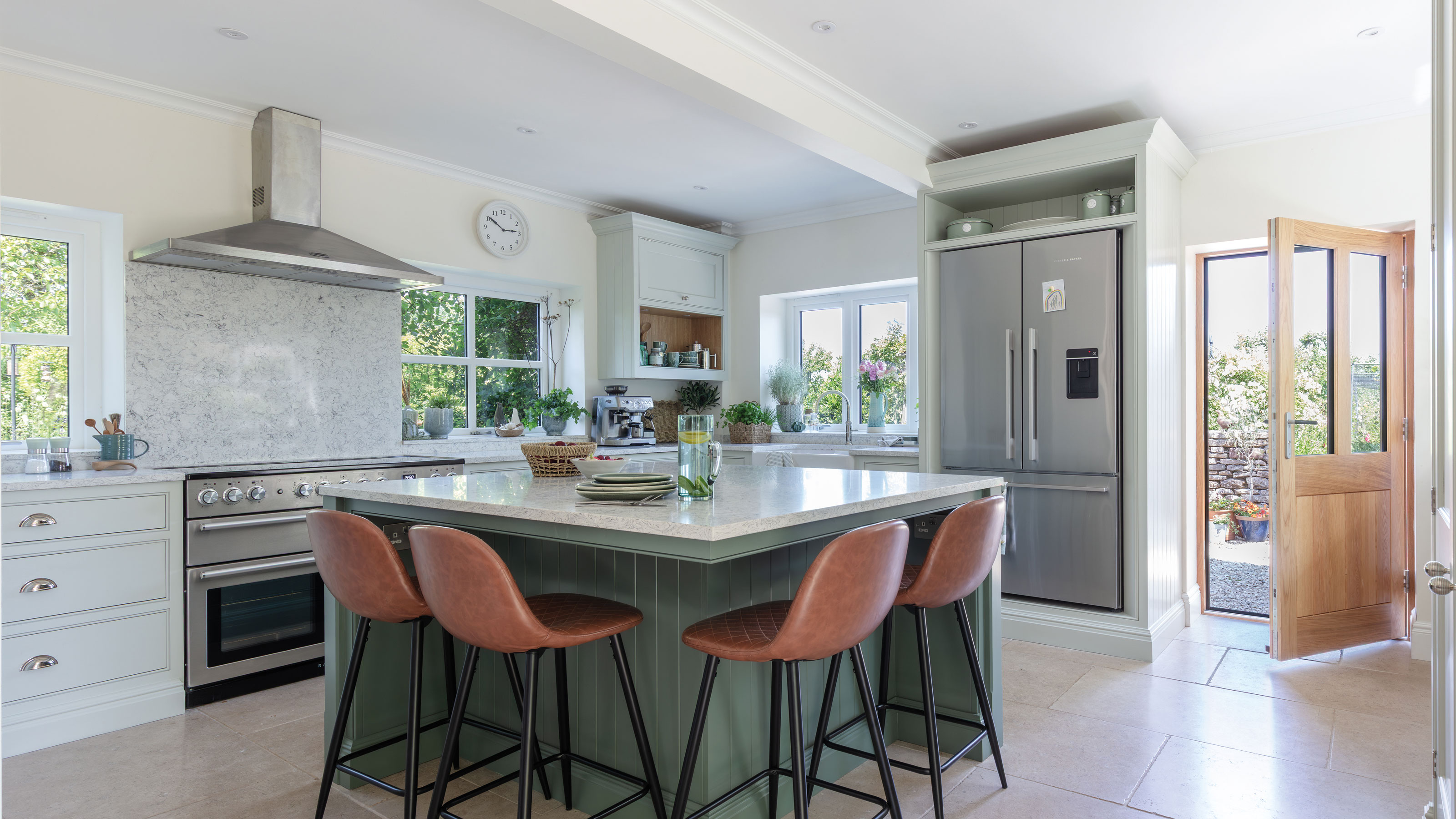 White kitchen with silver fridge and oven, a wooden door and green kitchen island in the foreground with stools around it