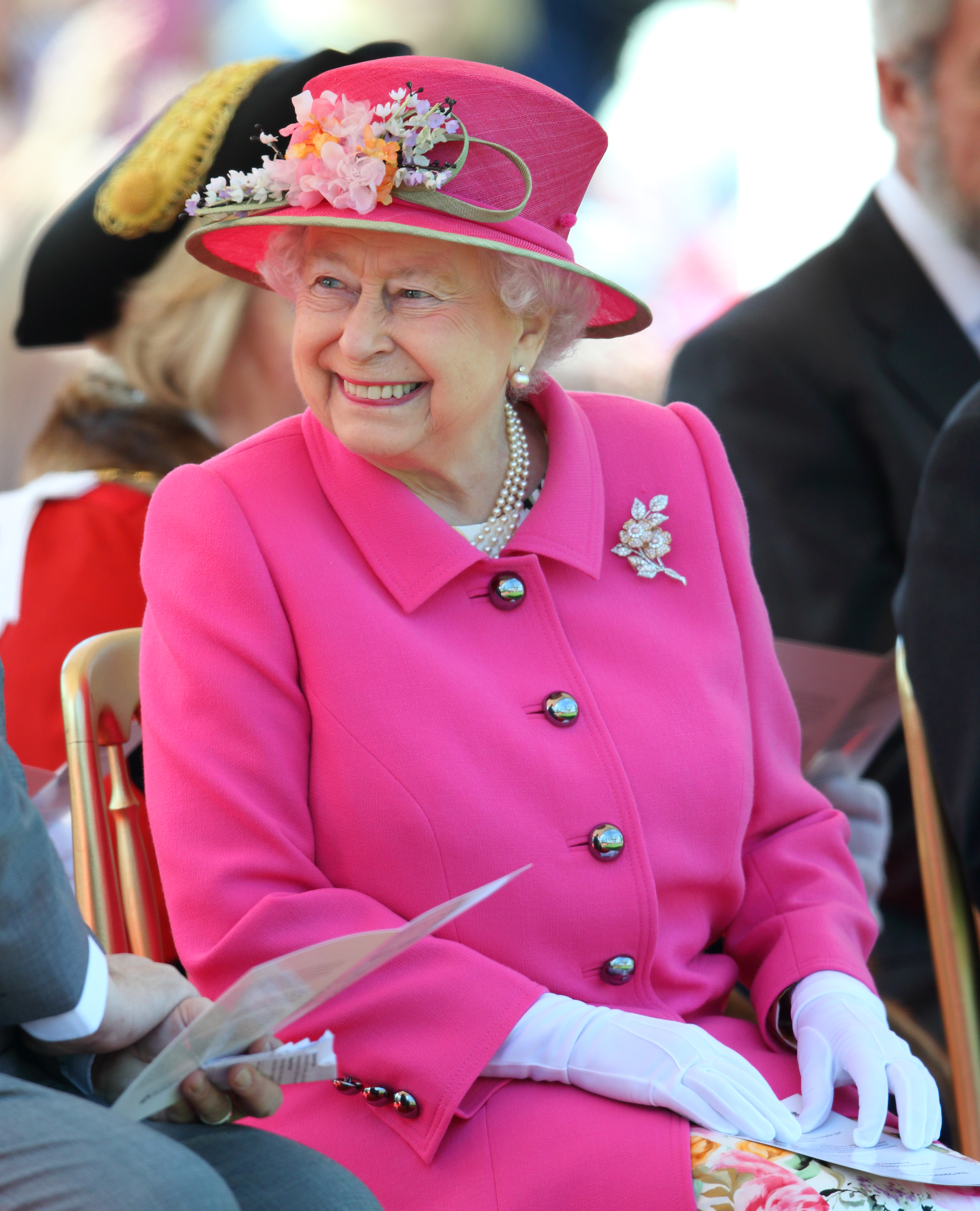 Queen Elizabeth wearing a pink coat and floral topped hat, sitting in a chair and laughing