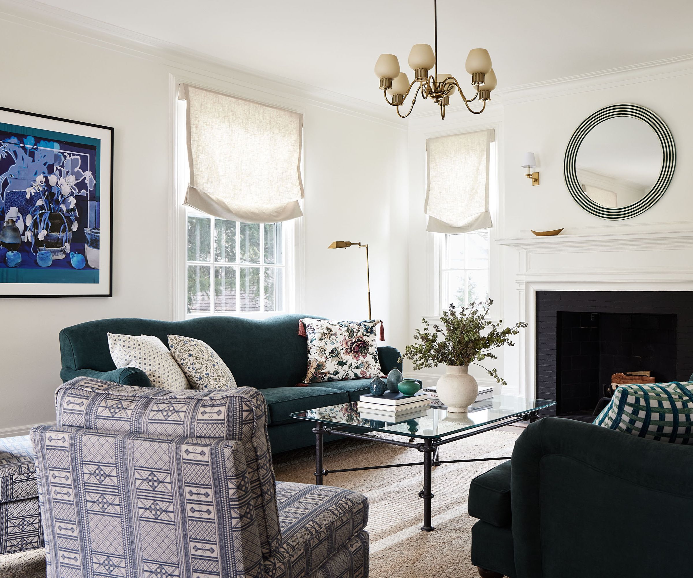 Blue and white living room with armchair and navy sofa, glass coffee table and fireplace with round mirror above it, and gold pendant light