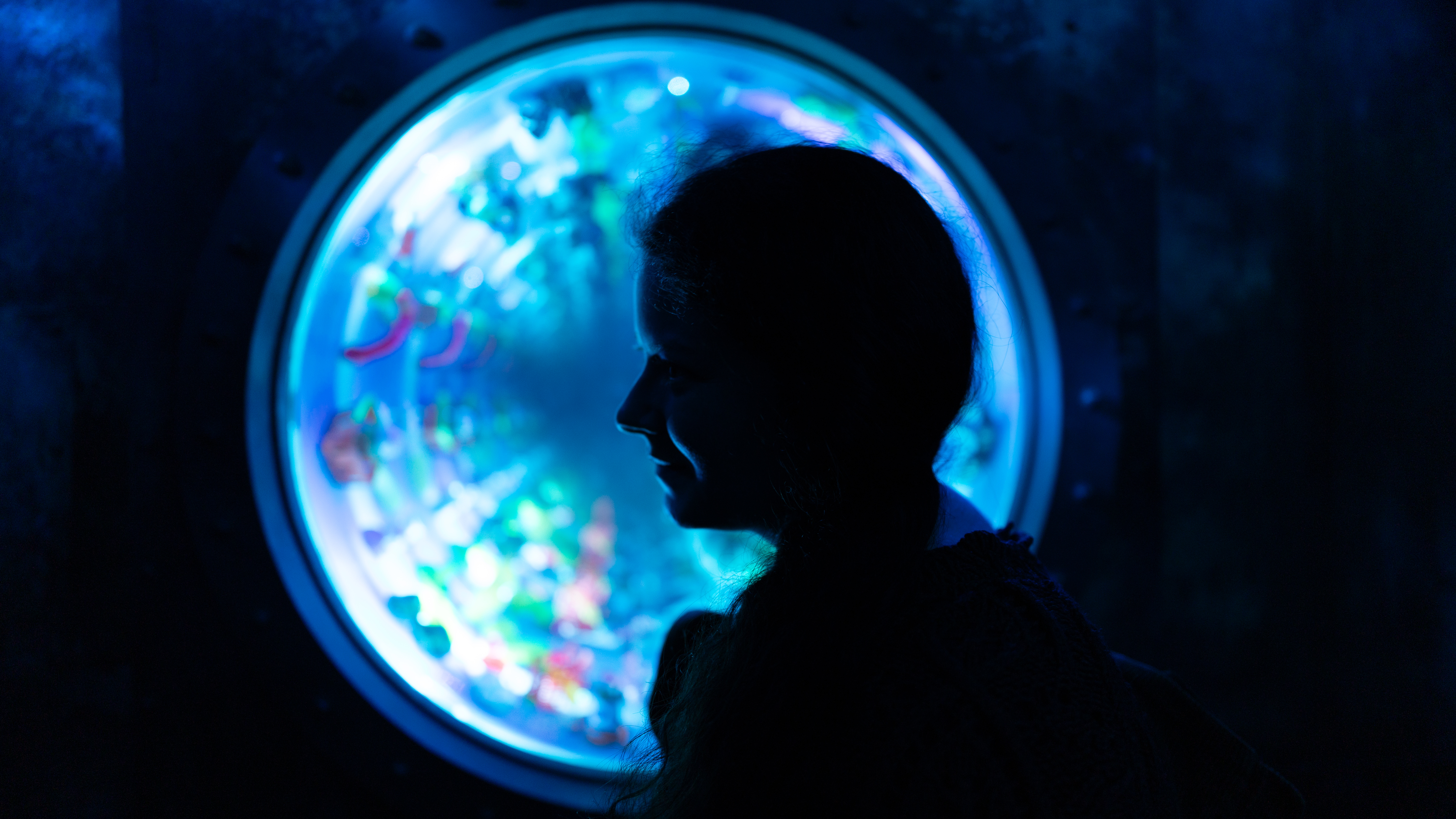 A low-light scene where a woman's face is backlit by a circular blue window with fluorescent lighting.