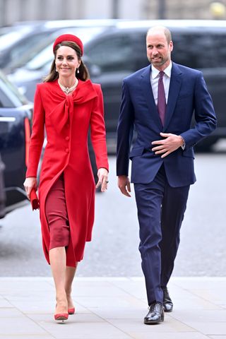 Kate Middleton wearing a red coat and headband walking outside next to Prince William in a blue suit