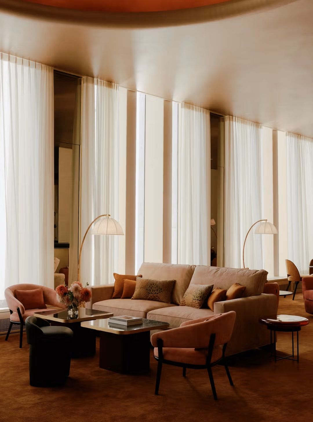 A 1970s-inspired lounge blends earthy, orangey tones with plush velvet seating, Art Deco standing lights in gold and glass, and flowy white curtains.