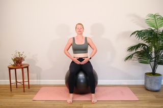 A woman sits on an exercise ball with her hands over her pelvis and her eyes closed as she performs pelvic floor exercises