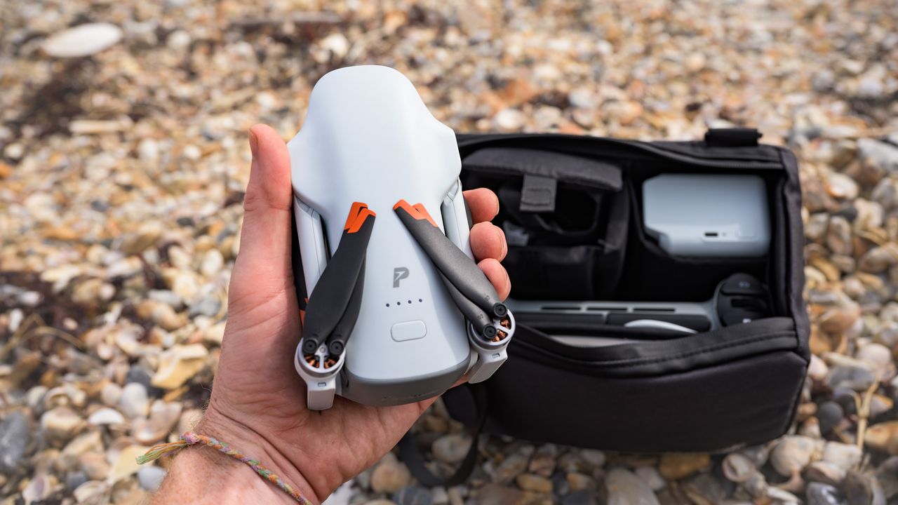 Potensic Atom 3 beginner drone in user's hands with its propellors folded away and the Fly More Combo bag and its content behind it, on a pebble beach