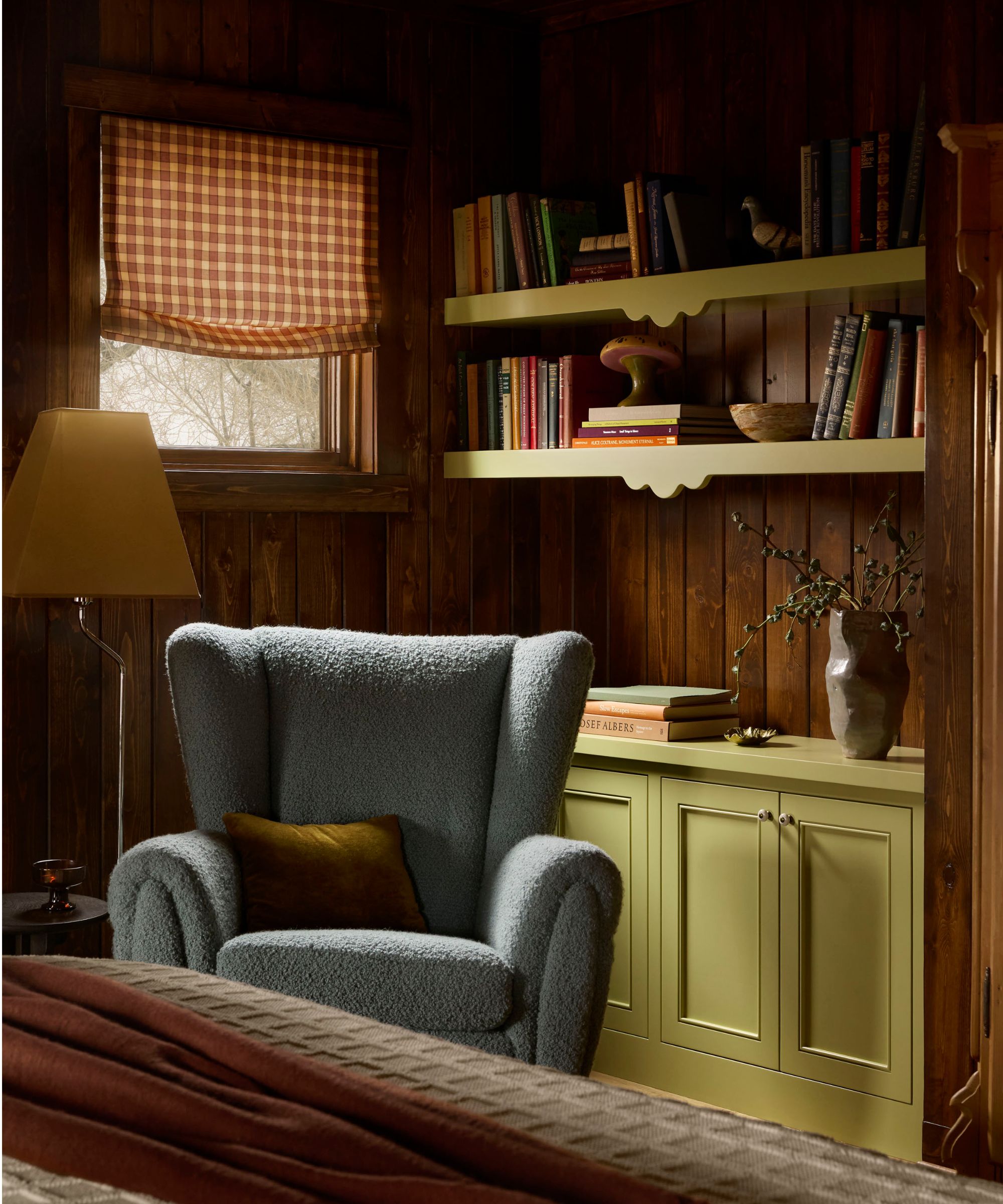 A bedroom in a cozy cabin with wood-clad walls, a small window with an ochre-patterned Roman blind, an alcove with cupboards and open shelving painted chartreuse with decorative objects and books, and a pale blue boucle armchair.