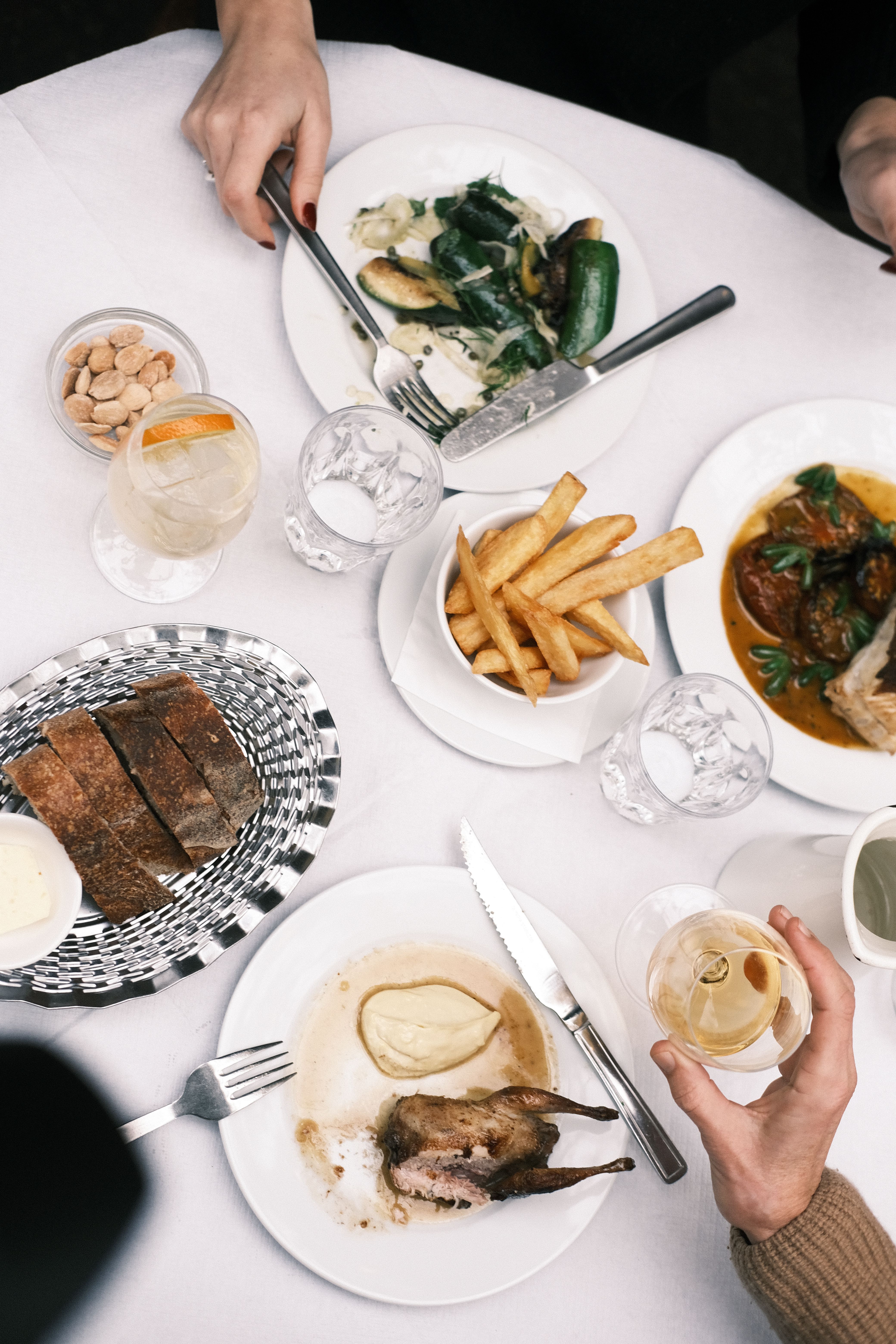 A busy, white-clothed dining table filled with delicious looking food and hands stretching to reach plates and wine glasses, seen from the above.