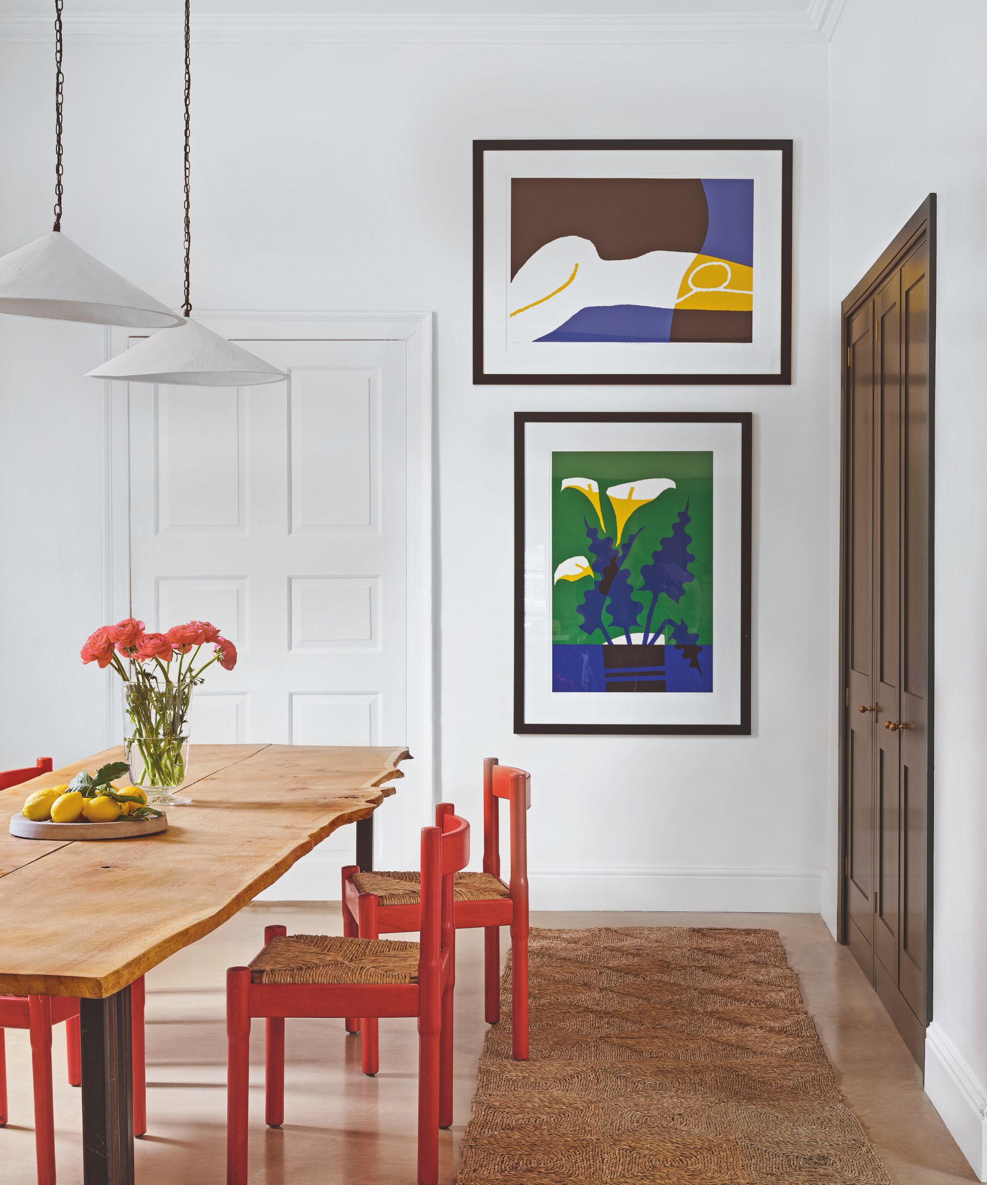 Dining room with white walls, a jute runner, an organic wood dining table, orange painted wooden dining chairs, white pendant lights above, and blue and green modern art hanging on the wall
