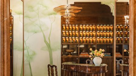 A whimsically decorated hotel lobby, with lacquered wooden cabinetry, a murano glass, flower-like chandelier, and handprinted tree murals in soft tones of green.