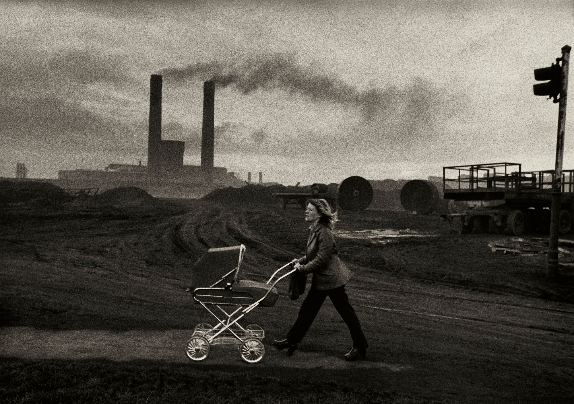 Black and white photograph of a woman pushing a pram along a bleak industrial road, with factory chimneys smoking in the background.