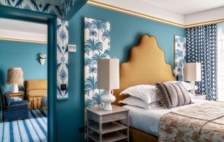 Blue bedroom and living room area with white able blue palm panels, stripy blue and white carpet and mustard headboard and couch