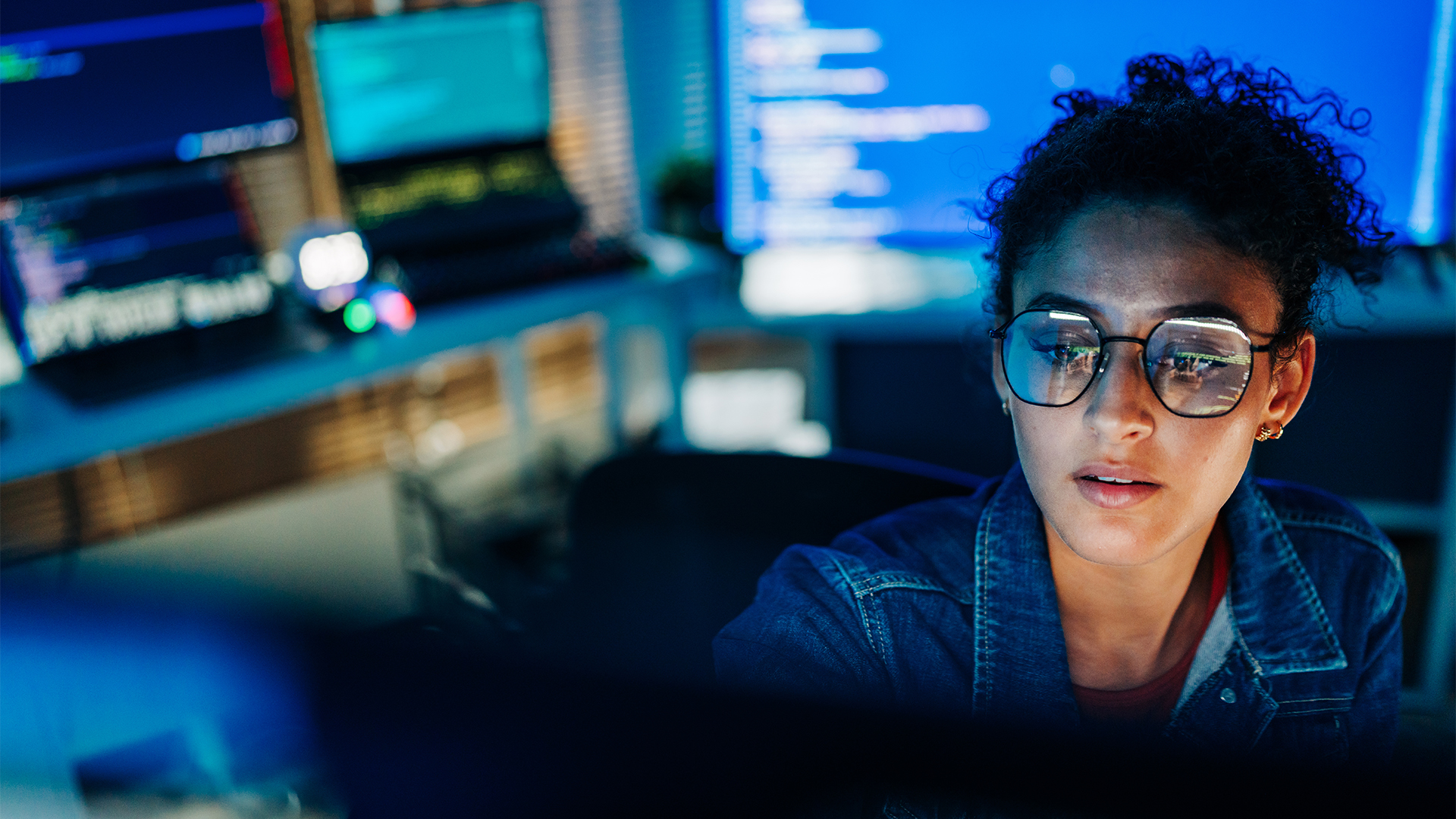Female software engineer with spectacles working on a desktop computer in a dimly-lit office space, with laptops showing computer code in background.