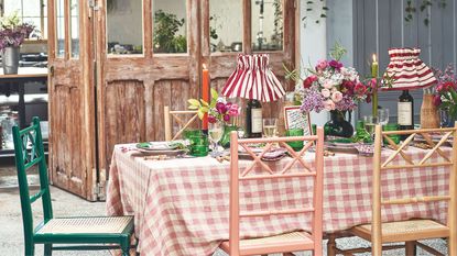 Rustic table with gingham tablecloth, rustic wooden colourful chairs, pleated striped lamps, taper candles, green glassware and flowers, with rustic salvaged doors in the background.