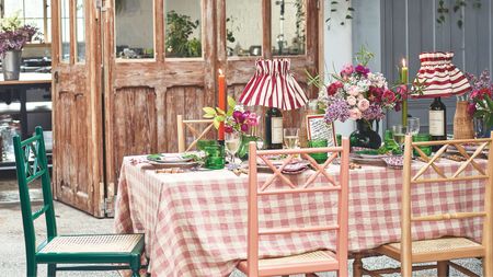 Rustic table with gingham tablecloth, rustic wooden colourful chairs, pleated striped lamps, taper candles, green glassware and flowers, with rustic salvaged doors in the background.