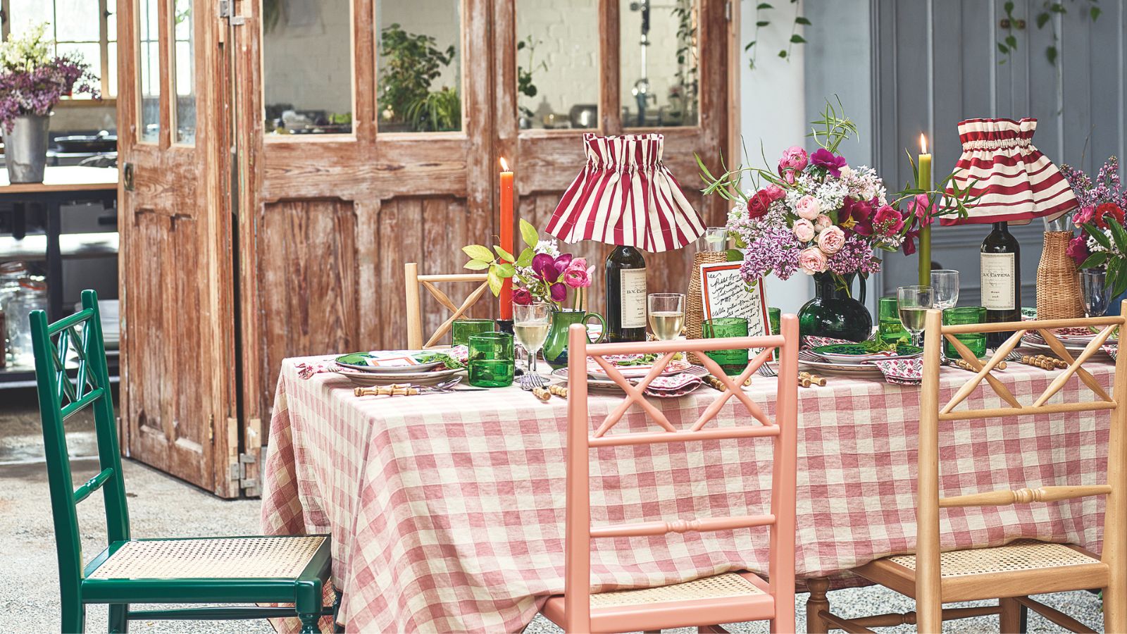 Rustic table with gingham tablecloth, rustic wooden colourful chairs, pleated striped lamps, taper candles, green glassware and flowers, with rustic salvaged doors in the background.