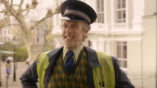 Peter Capaldi stands grinning in a doorway wearing his safety vest and hat in Paddington 2.