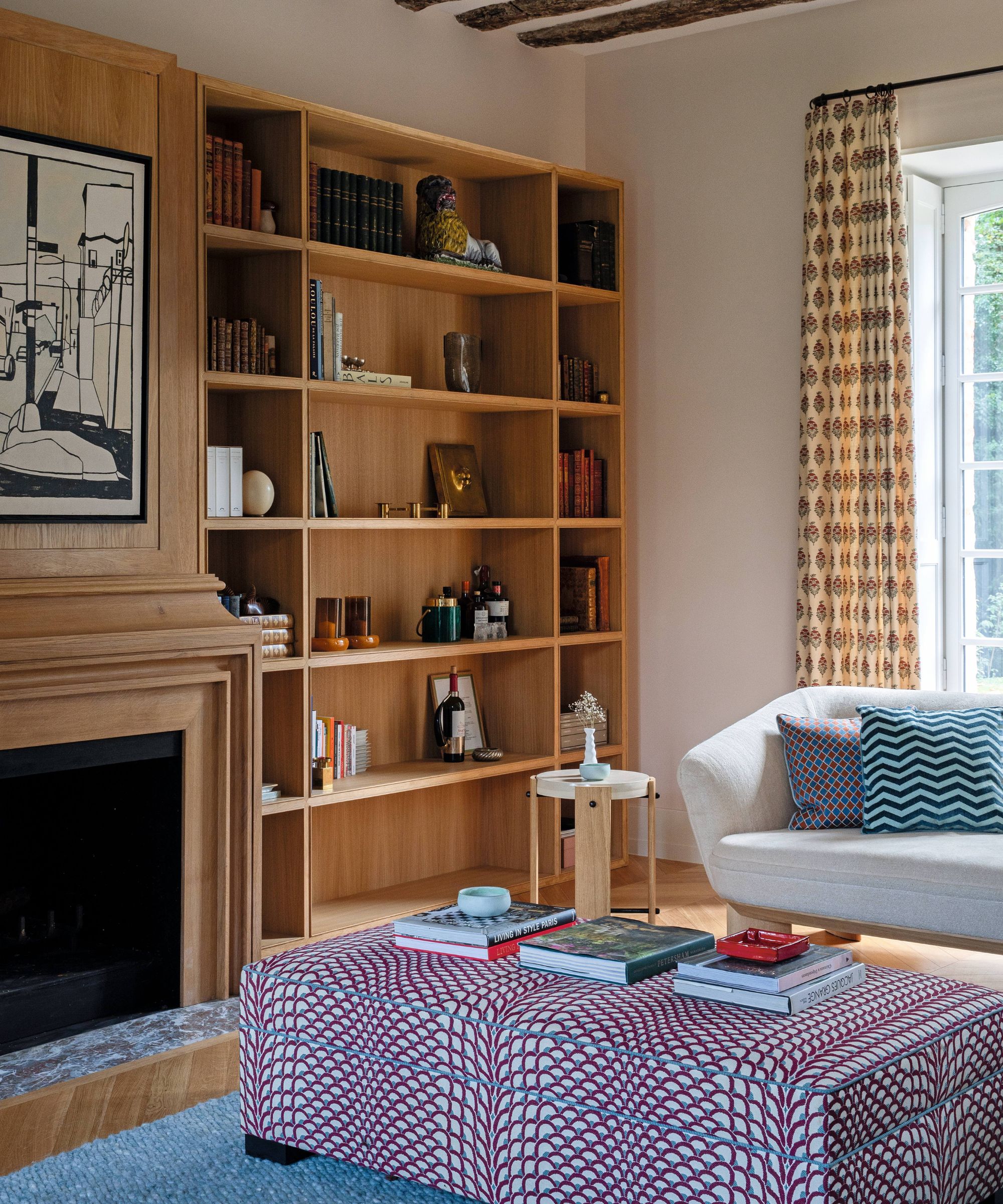 a sitting room in a french chateau with oak bookcases and fireplace, rustic ceiling beams, a white sofa, and patterned ottoman