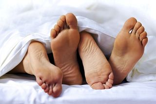 A couples feet stick out of the sheets at the end of a bed.