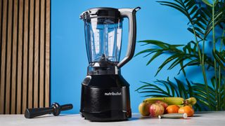 a black nutribullet with a big blending component is photographed in front of a blue backdrop, showing its plastic pitcher jug, silver blades, and tamper