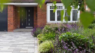 block paved driveway leading to brick house with modern grey front door, garage door and heavily planted area in front of house