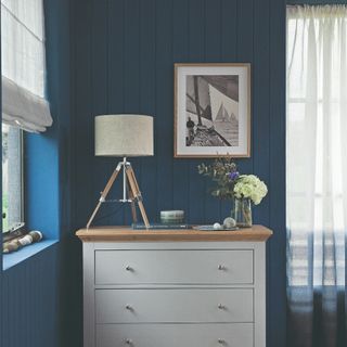 A room covered in blue tongue and groove wall panelling contrasted by a grey chest of drawers