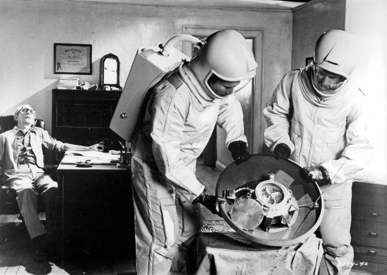 grey-scale image with a man asleep in a chair at a desk against the wall in the background, as two haz-mat suited men handle some sort of circular mechanical equipment.