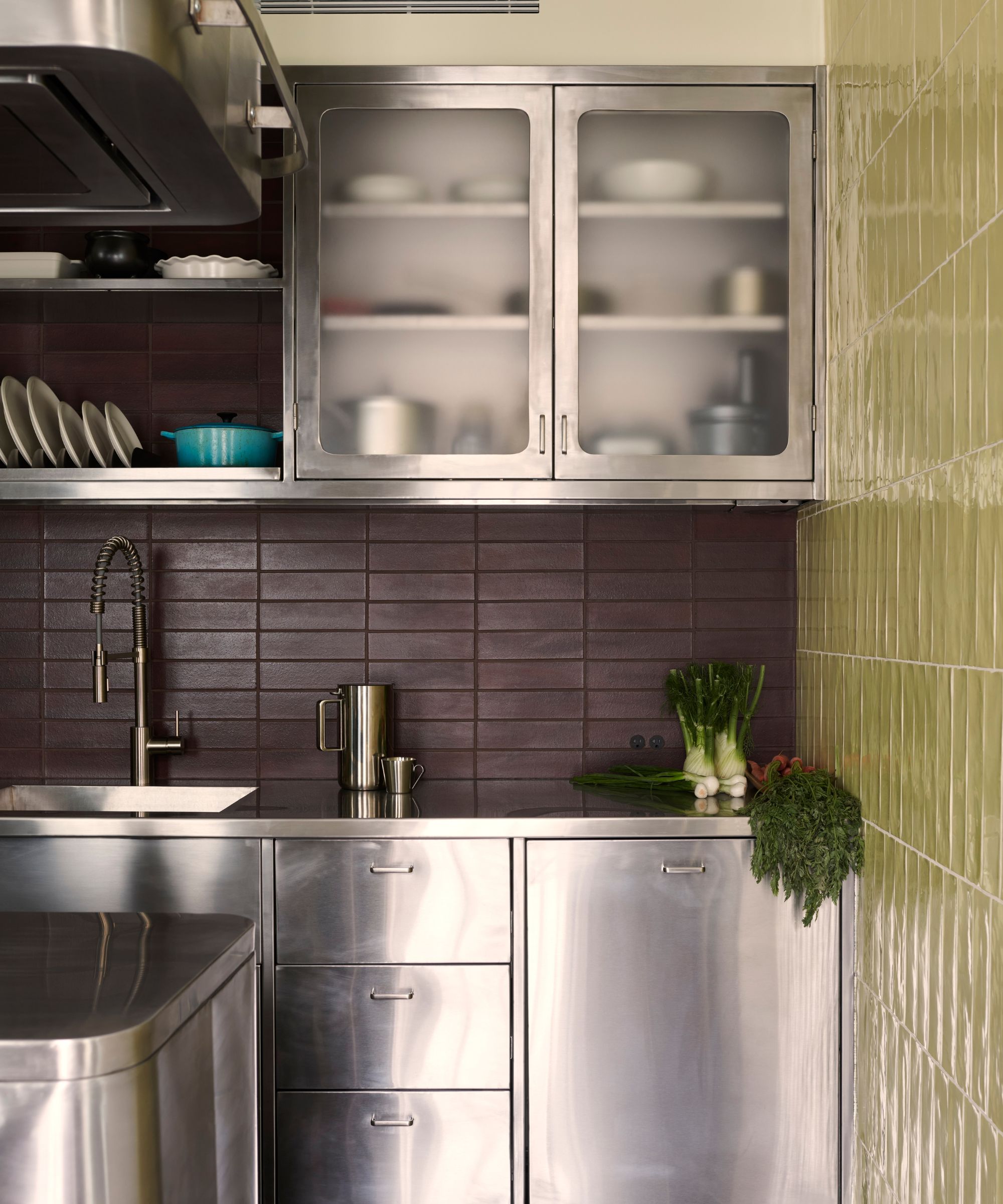 A professional-grade kitchen station with stainless steel lower cabinets and frosted glass upper cabinets. The walls are finished in a mix of deep burgundy and sage green subway tiles, paired with a commercial-style tall faucet.