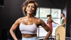 A Pilates instructor stands in a studio with her hands on her hips, looking at the camera.
